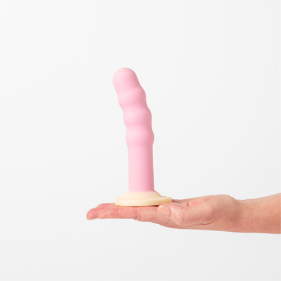 Hand holding Dilly, Girls Get Off's pink, ribbed silicone dildo with a cream suction base against a white background.