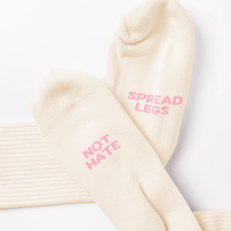 A pair of nude color crew socks with pink phrases of "Spread Legs" and "Not Hate"