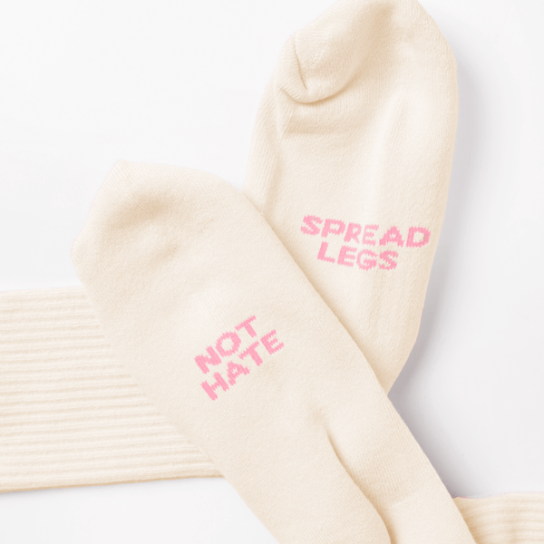 A pair of nude color crew socks with pink phrases of "Spread Legs" and "Not Hate"