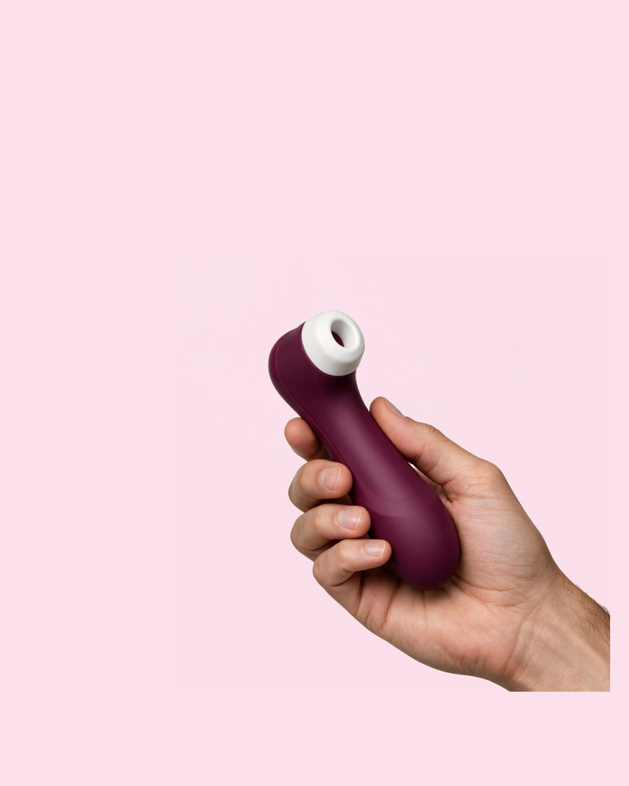 A hand holding a wine red Satisfyer Pro 2 air pulse vibrator against a plain background.