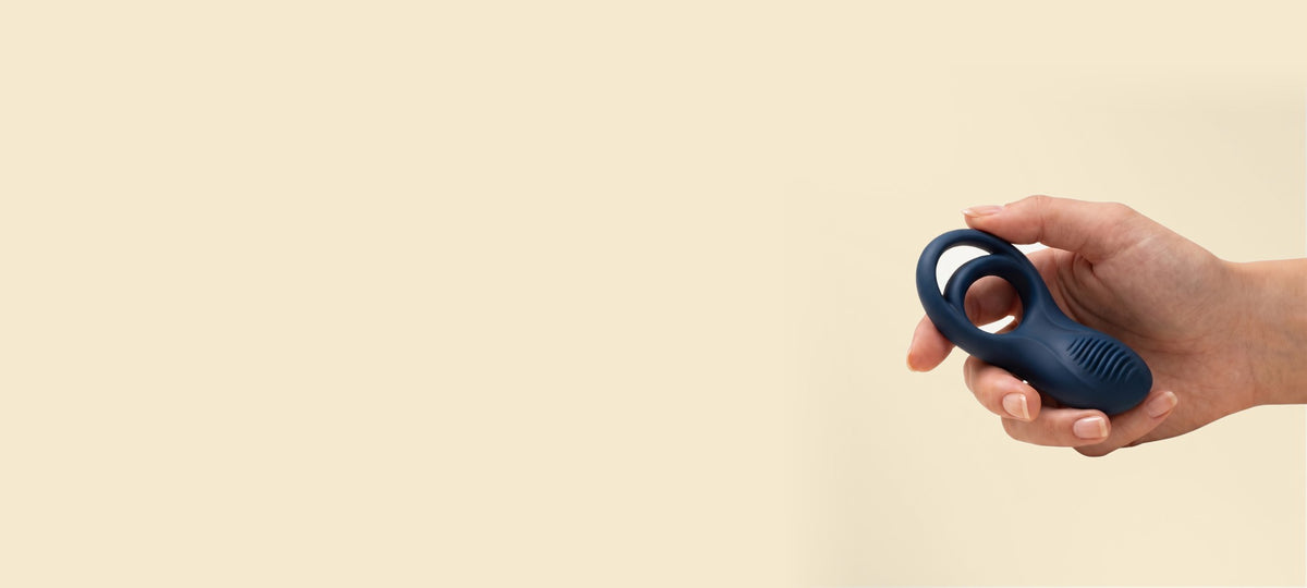 A hand holding the navy Satisfyer Epic Duo cock and ball ring against a plain cream background