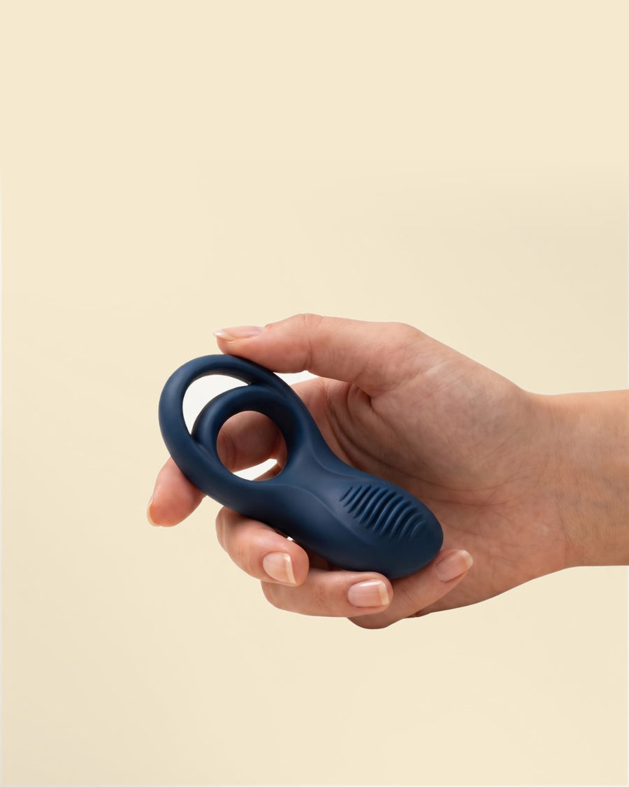 A hand holding the navy Satisfyer Epic Duo cock and ball ring against a plain cream background